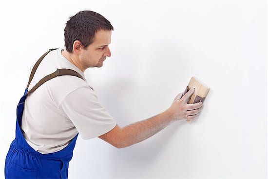 a man in royal blue pants engaging in surface preparation (sanding a wall), getting ready for painting